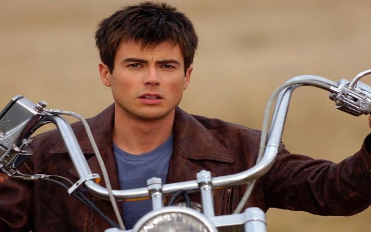 Matt Long in a dark brown leather jacket and blue t-shirt riding a bike.